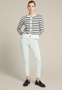 Schwarz-weißer gestreifter Cardigan mit rundem Ausschnitt, langen Ärmeln und Knopfverschluss. Kombiniert mit hellmintgrünen Slim-Fit-Hosen und schwarzen Schuhen.