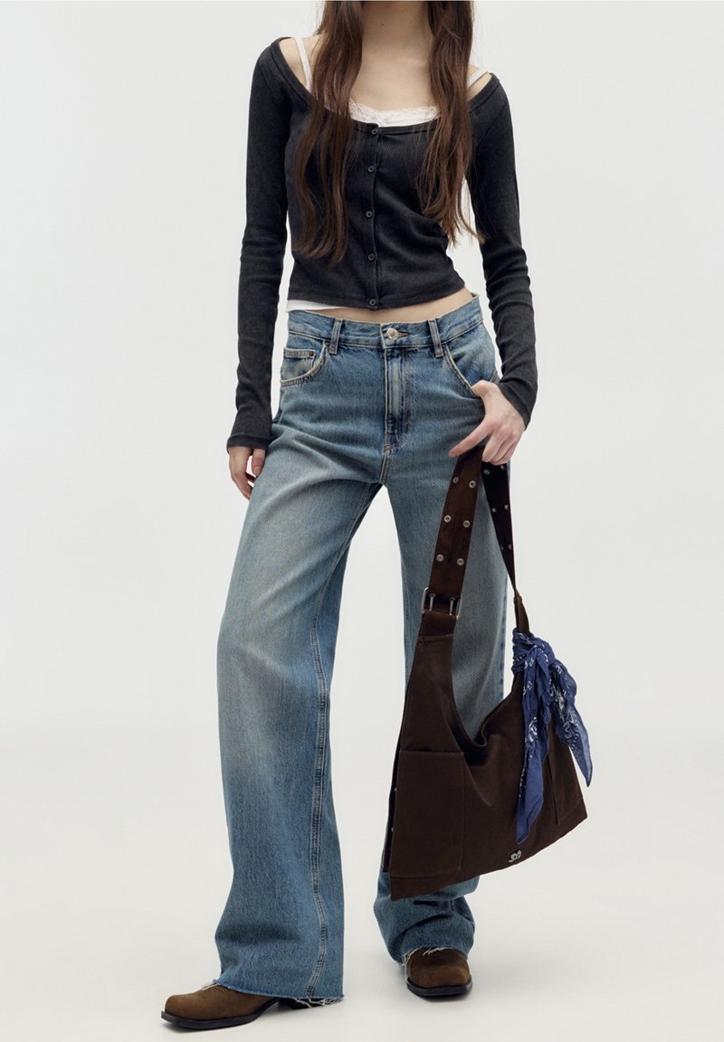 Jeune femme portant un haut noir à manches longues et un jean large, tenant un sac à épaule marron avec un bandana bleu attaché.
