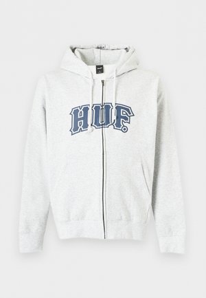 Jeune homme portant un sweat à capuche zippé gris clair avec le logo "HUF" en lettres foncées, associé à un jean bleu foncé sur fond blanc uni.