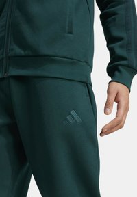 Tuta sportiva verde scuro con cerniera. Il materiale è liscio con polsini a costine. Presenta un logo delicatamente in rilievo sui pantaloni.