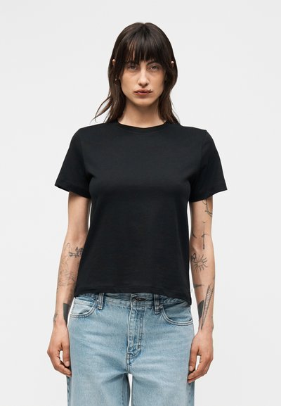 Kvinne med mørkt hår og tatoveringer på begge armer iført en ensfarget svart kortermet t-skjorte og lyseblå jeans foran en hvit bakgrunn.