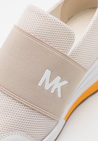 Baskets à enfiler en mesh beige et blanc avec une large bande élastique sur le dessus, ornée du logo "MK" blanc en relief et d'un accent orange sur la semelle.