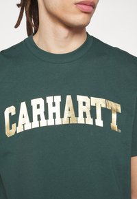 Osoba w ciemnozielonej koszulce z dużym, metalicznym, złotym napisem "CARHARTT" na piersi, skrócona poniżej nosa.