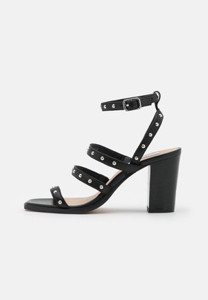 Sandale noire à talon haut avec quatre brides cloutées et une boucle de cheville réglable sur fond blanc.