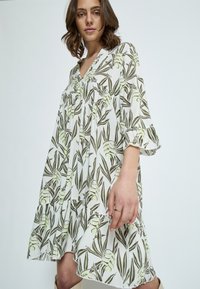 Weißes Kleid mit grünem und braunem Blätterdruck, 3/4-Ärmel, gestuftem Rock und V-Ausschnitt. Hergestellt aus leichtem Stoff.
