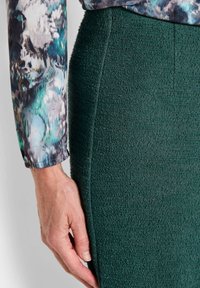 Strapazierfähiger grüner Rock mit tailliertem Schnitt, kombiniert mit einem Langarmshirt mit einem mehrfarbigen Blumenmuster. Eine Hand ruht auf dem Rock.