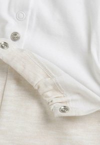 Tissu en coton blanc avec des fermetures à pression se superpose à un matériau beige texturé, montrant un ourlet roulé et des détails de couture soignés.