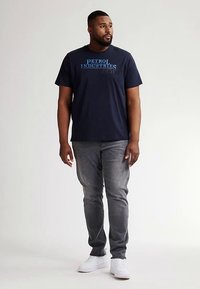 Tmavě modré bavlněné tričko s modrým potiskem "PETROL INDUSTRIES", doplněné šedými džíny a bílými teniskami. Model stojí vzpřímeně na neutrálním pozadí.