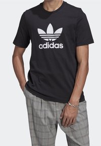Černé bavlněné tričko s velkým bílým logem trefoil Adidas a textem, kulatým výstřihem a krátkými rukávy. Volný střih s viditelným švěm.