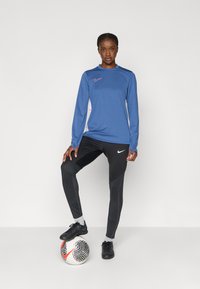 Μπλε αθλητική μπλούζα με μακριά μανίκια και ψηλό λαιμό, συνδυασμένη με μαύρο εφαρμοστό παντελόνι με υφασμάτινα σχέδια και μαύρα ποδοσφαιρικά παπούτσια.