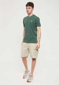 T-shirt verde in cotone, colletto rotondo, maniche corte, abbinato a pantaloni cargo beige, con tasche laterali. Sneakers grigio chiaro con dettagli in marrone.