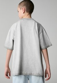 Šedé oversized tričko s krátkým rukávem a kulatým výstřihem, s měkkou texturou a volným střihem, částečně zasunuté do světlých džínů.