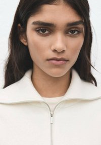 Približen posnetek osebe s temnimi lasmi, oblečene v belo zgornjo obleko z zadrgo in visokim ovratnikom; tkanina je gladka s subtilno teksturo.