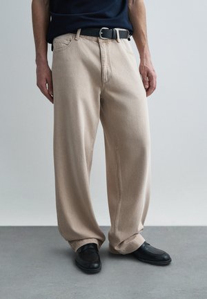 Μπεζ τζιν παντελόνια φαρδιάς γραμμής, υφασμάτινη υφή, μπροστινές τσέπες, θηλιές για ζώνη, φορεμένα με μαύρα παπούτσια, μινιμαλιστικός σχεδιασμός, ουδέτερη χρωματική παλέτα.