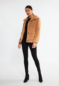 Tan faux-shearling jakke med krave, to forlommer og sorte knapper. Båret over en sort top og leggings, med sorte ankelstøvler.