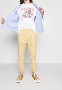 T-shirt blanc avec un graphisme coloré de sel et vinaigre, chemise boutonnée bleu clair et pantalon jaune. Des baskets montantes marron complètent le look.