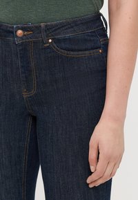 Jeans in denim blu scuro con una vestibilità classica, hardware in tono rame e dettagli di cucitura sottili. Mano appoggiata sulla coscia.