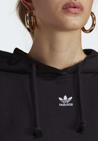 Schwarzer Kapuzensweater aus weichem Material, mit einem weißen, auf die Brust gestickten Adidas-Logo und Zugbändern, die in Pompons enden.