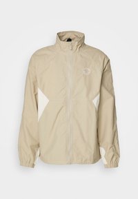 Chaqueta ligera beige con cremallera frontal completa, cuello alzado, puños elásticos, paneles laterales blancos y un pequeño logo en el pecho.