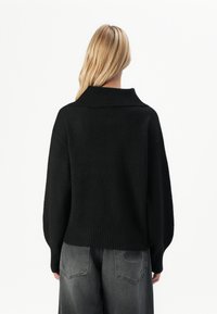 Svart, strikket genser med bred, ribbestrikket krage og lange, løse ermer. Brukt sammen med grå, lett slitte bukser med et logopatch.