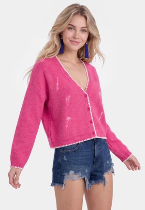 Jeune femme portant un cardigan rose boutonné avec une broderie florale blanche et un short en jean bleu déchiré, souriante avec de longs cheveux blonds.