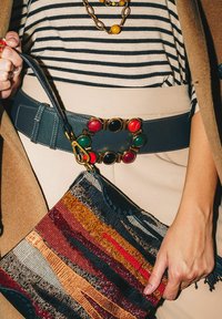 Borsa multicolore con trama intricata, caratterizzata da strisce orizzontali in rosso, oro e blu navy. Ampia cintura blu con accenti di pietre colorate.