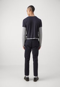 Un t-shirt bleu marine à manches courtes superposé sur une chemise à rayures à manches longues, associé à un pantalon sombre et des chaussures noires, vu de l'arrière.