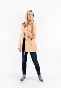 Jakna u boji breskve otporna na vodu s kapuljačom, podesivim špagama, džepovima na zip i detaljima od srebra, nosi se uz tamne traperice i tenisice.