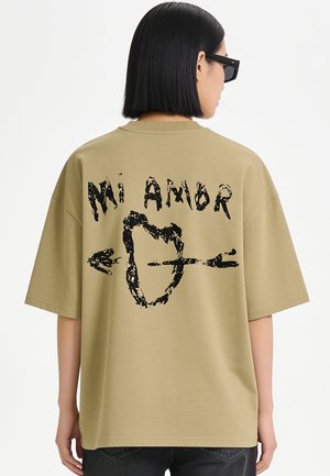 Personne avec cheveux courts noirs portant des lunettes de soleil et un t-shirt oversize vert olive avec un texte noir "Mi Amor" et un graphisme de cœur avec une flèche.