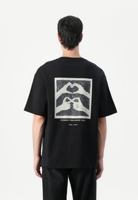 Black cotton t-shirt with short sleeves featuring a graphic print of hands forming a heart and connecting, text reads "CONNECTING SINCE 1993."