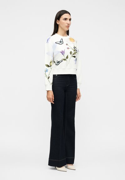 Γυναίκα που φοράει άσπρο φούτερ με σχέδια πεταλούδας και λουλουδιών, σκούρα καμπανέ τζιν και άσπρα μυτερά παπούτσια, στέκεται μπροστά σε απλό φόντο.