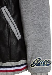 Sort læder og grå stofjakkeærme med Guess-logo patch og stribet ribkantsmanchet i rød, hvid og marineblå.