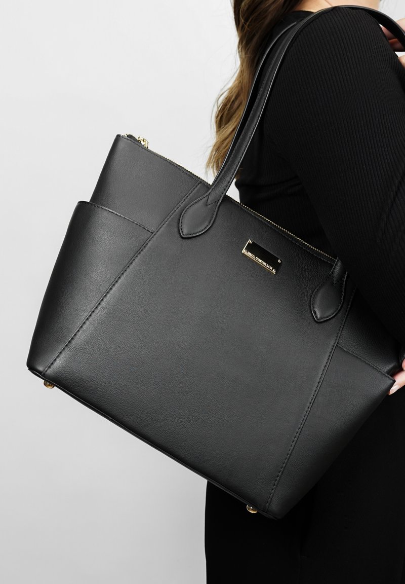 Sac fourre-tout en cuir noir avec des poignées robustes, une forme structurée, des détails en métal doré et une fermeture zippée. Dispose d'une poche latérale pour un rangement supplémentaire.