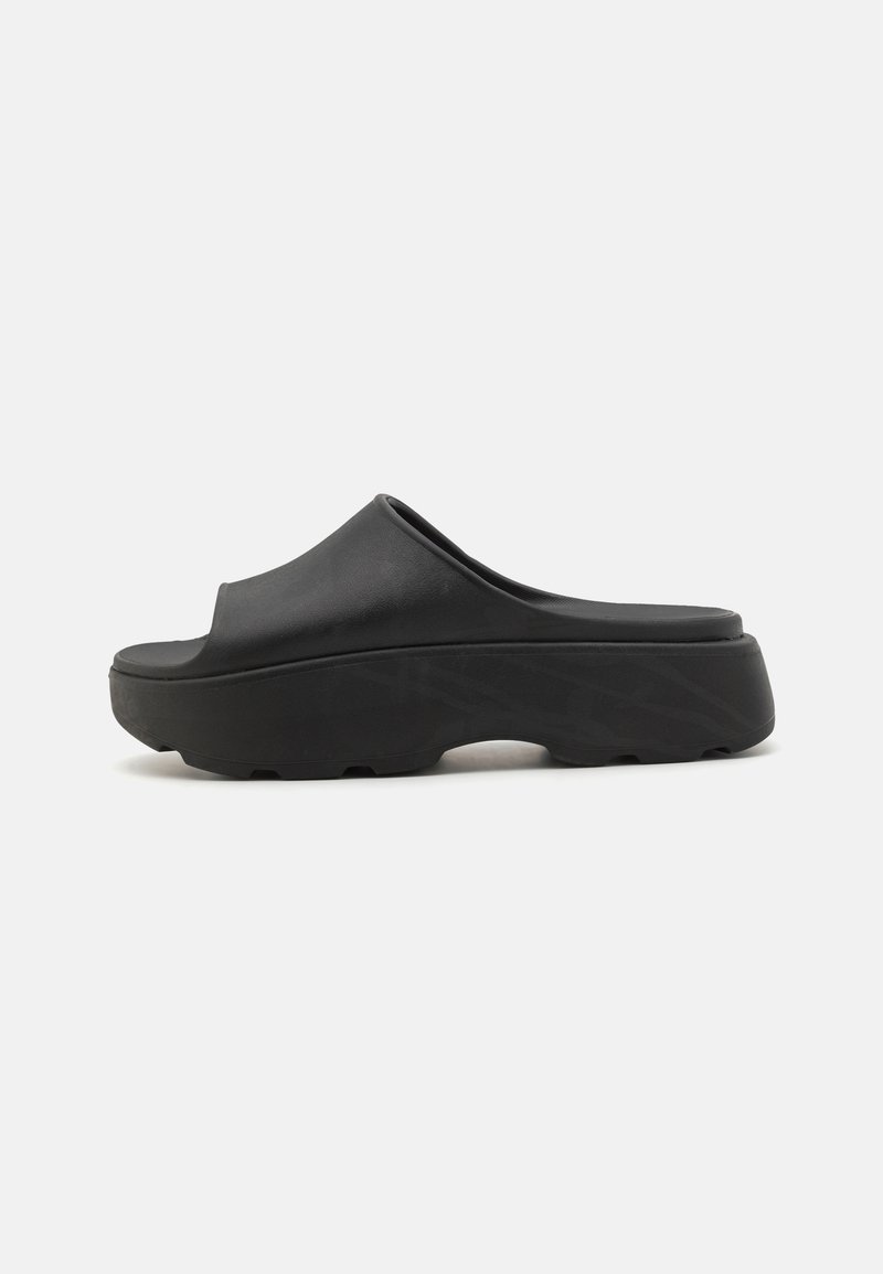 Svarte slip-on klosser med bred åpen overdel, tykk teksturert såle og formet fotbed. Materialet ser fleksibelt og holdbart ut.