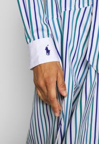 Striped button-up shirt with blue, green, and white stripes. Features a white cuff with a blue embroidered logo of a horse and rider.