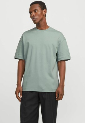 Jeune homme aux cheveux courts portant un T-shirt ample vert sauge et un pantalon noir, se tenant devant un fond clair uni.