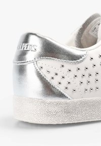 Witte leren sneaker met zilveren metallic accenten, versierd met kleine studs. Getextureerde witte zool met een subtiel patroon en een afgeronde hak.