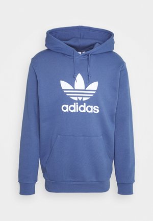 Sininen huppari, joka on valmistettu puuvillasekoitekankaasta. Hupparissa on valkoinen adidas-logo ja graafinen kuva kolmesta raidasta. Siinä on etutasku ja nyörit.