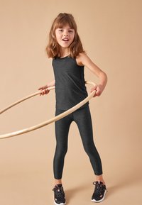 Temno siva športna majica brez rokavov in pajkice, združene s črnimi supergami. Otrok drži lesen hula hoop, na nevtralni podlagi.