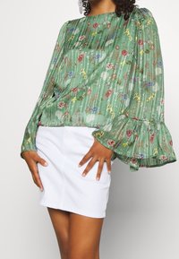 Blusa in chiffon floreale verde con maniche lunghe e volant, abbinata a una gonna di jeans bianca. Presenta un design testurizzato con fiori di vari colori.
