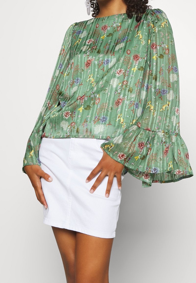 Blusa in chiffon floreale verde con maniche lunghe e volant, abbinata a una gonna di jeans bianca. Presenta un design testurizzato con fiori di vari colori.