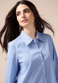 Camisa azul de botones con rayas verticales blancas y doradas. Estilo de cuello, mangas largas, con un brillo sutil, combinada con pendientes ovalados plateados.