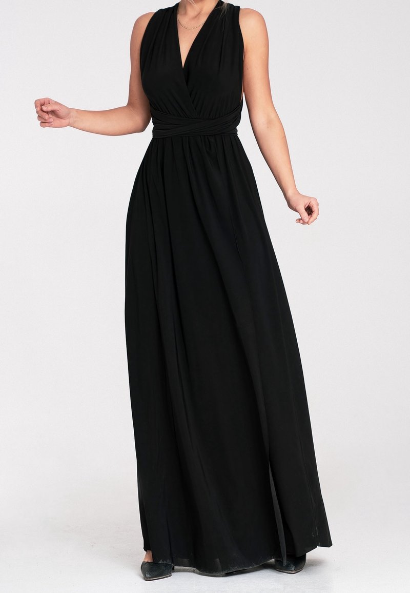 Femme portant une robe maxi noire sans manches avec un profond décolleté en V et une taille froncée, debout devant un fond clair uni.
