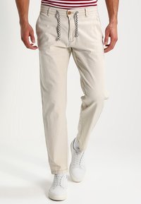 Hombre que lleva pantalones casuales beige con cordones en blanco y negro, zapatillas blancas y una camiseta a rayas rojas y blancas, visto de la cintura hacia abajo.
