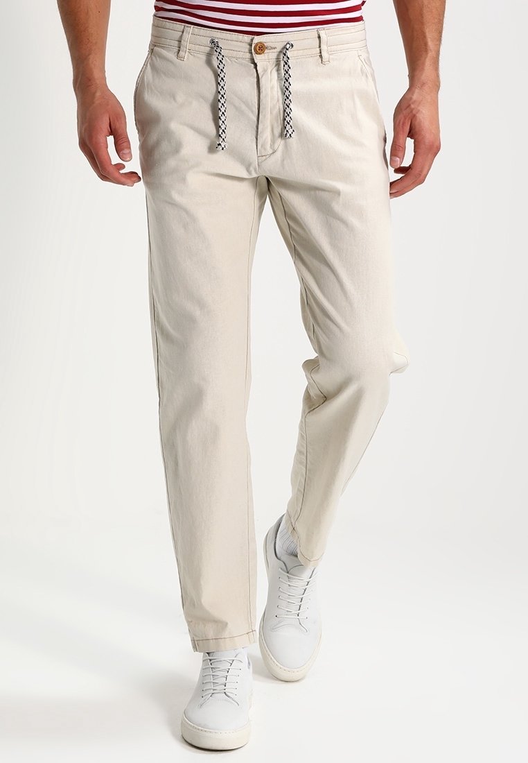 Hombre que lleva pantalones casuales beige con cordones en blanco y negro, zapatillas blancas y una camiseta a rayas rojas y blancas, visto de la cintura hacia abajo.