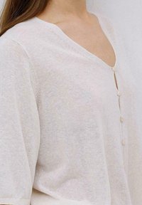 Helles Creme, strukturiertes Stricktop mit V-Ausschnitt, kurzen Ärmeln und Knopfleiste. Verfügt über zarte Knöpfe und eine lockere Passform.