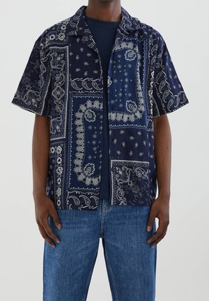 Mężczyzna noszący granatową koszulę z krótkim rękawem z białymi wzorami paisley i kwiatowymi na bandanie, na ciemnym t-shircie i niebieskich dżinsach.