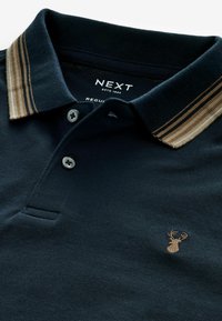 Mörkblå polo skjorta i texturerad tyg, med en kontrasterande beige randig krage och en liten broderad älglogotyp på bröstet. Tvåknappsstängning.