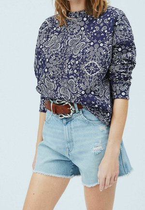 Woman wearing a navy blue paisley long-sleeve top, distressed light denim shorts, and a brown leather belt with a unique silver buckle.