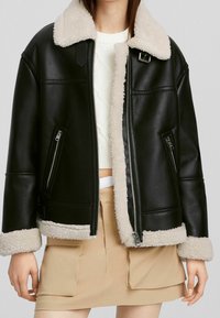 Kvinna som bär en svart konstläderjacka med krämfärgad teddykrage, manschetter och kant, ihop med en vit stickad tröja och en beige kjol med fickor.